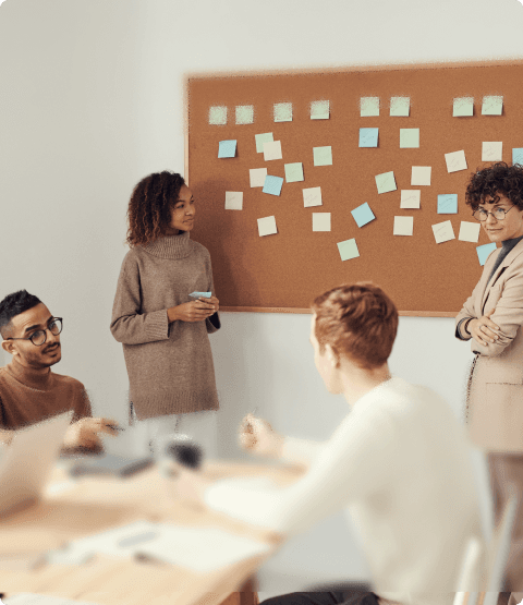 A close-up of a person's hand holding a post-it and presenting ideas in front of other people.